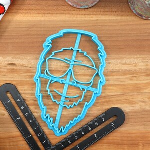 May include: A blue plastic cookie cutter shaped like a person with a beard and sunglasses. The cookie cutter is on a wooden surface.