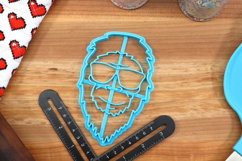 May include: A blue plastic cookie cutter shaped like a person with a beard and sunglasses. The cookie cutter is on a wooden surface.