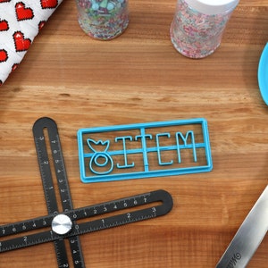 May include: Blue plastic cookie cutter with the word "ITEM" and a small donut design. The cutter is on a wooden surface with a black ruler, a rolling pin, a blue spatula, and two jars of sprinkles.