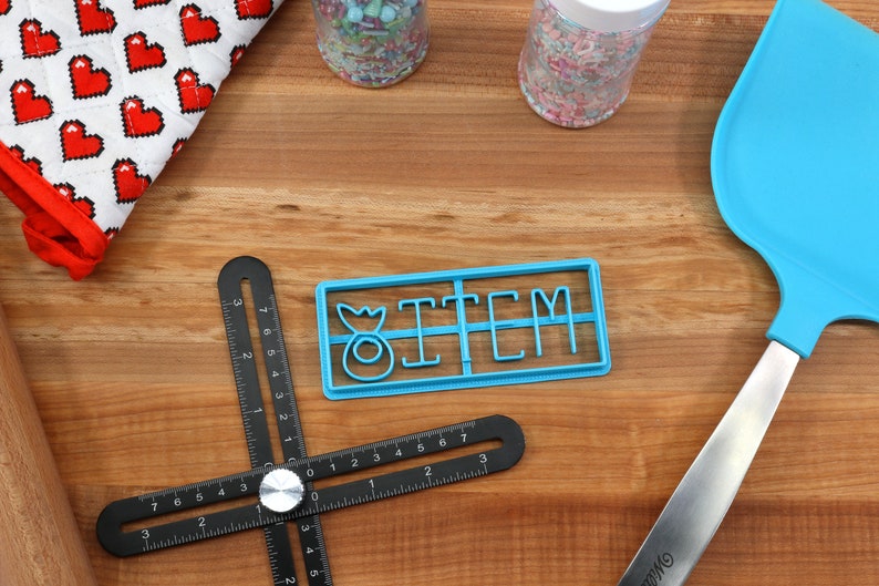 May include: Blue plastic cookie cutter with the word "ITEM" and a small donut design. The cutter is on a wooden surface with a black ruler, a rolling pin, a blue spatula, and two jars of sprinkles.