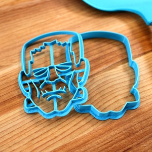 May include: Two blue plastic cookie cutters shaped like Frankenstein's face. The cutters are on a wooden surface.
