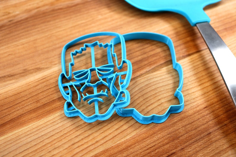 May include: Two blue plastic cookie cutters shaped like Frankenstein's face. The cutters are on a wooden surface.
