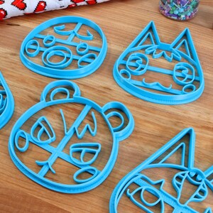 May include: Four blue plastic cookie cutters shaped like animal faces. The cutters are on a wooden surface.
