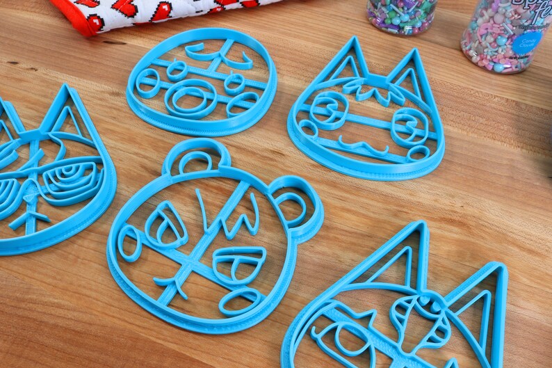 May include: Four blue plastic cookie cutters shaped like animal faces. The cutters are on a wooden surface.