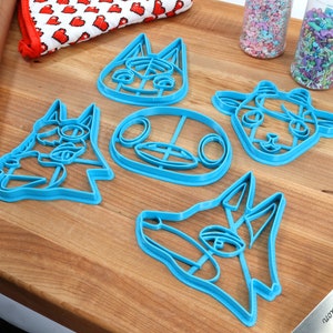 May include: Set of five blue plastic cookie cutters shaped like animal faces. The cutters are on a wooden surface.