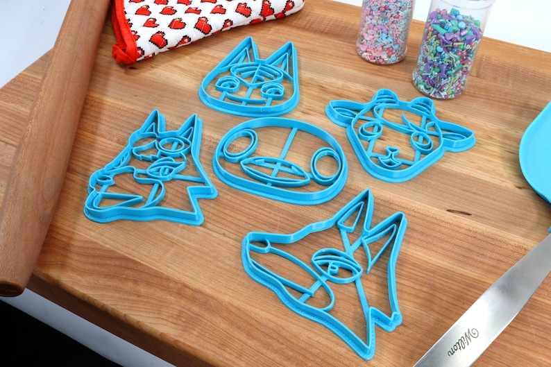 May include: Set of five blue plastic cookie cutters shaped like animal faces. The cutters are on a wooden surface.