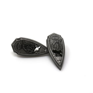 May include: Two silver-colored metal shields with a dragon design. The shields are shaped like a teardrop and have a raised edge.