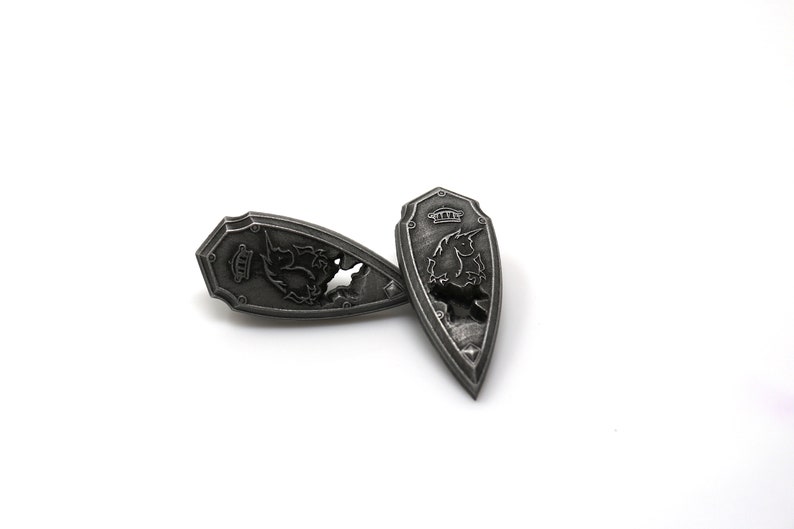 May include: Two silver-colored metal shields with a dragon design. The shields are shaped like a teardrop and have a raised edge.