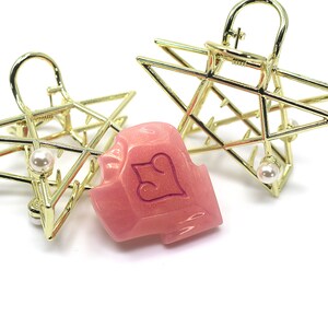 May include: Two gold-colored star-shaped hair clips with a pearl accent and a pink hair clip with a heart design. The hair accessories are on a white background.