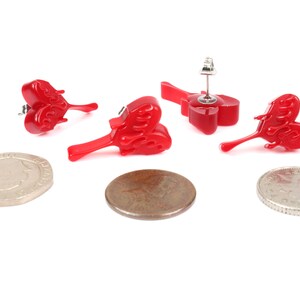 May include: Three pairs of red heart-shaped earrings with a flame design. The earrings are shown on a white background with two coins.