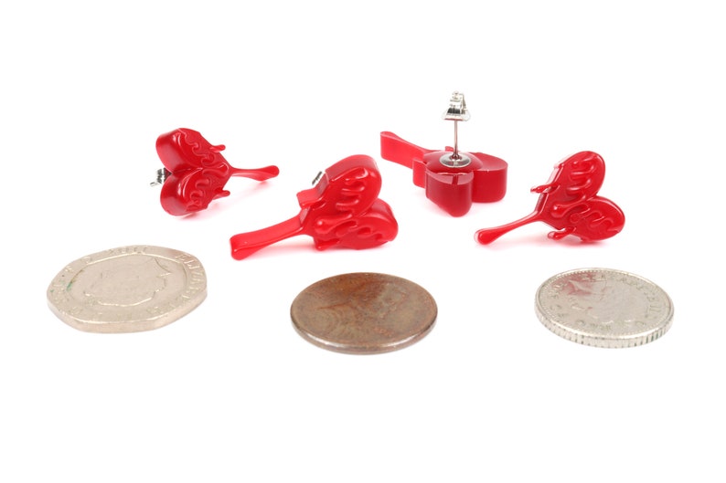May include: Three pairs of red heart-shaped earrings with a flame design. The earrings are shown on a white background with two coins.