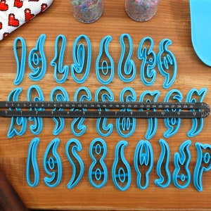 May include: A set of blue plastic cookie cutters shaped like the numbers 1 through 9. The cutters are arranged in three rows on a wooden surface. A ruler is placed between the first and second rows.