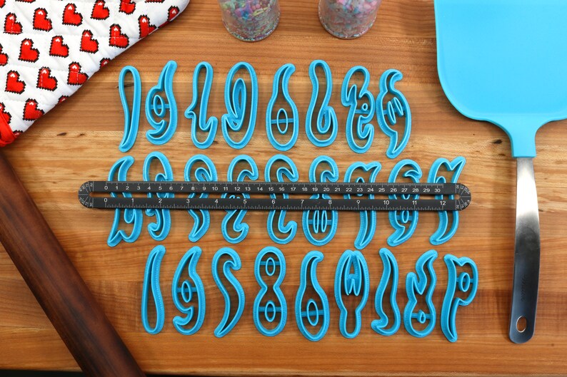 May include: A set of blue plastic cookie cutters shaped like the numbers 1 through 9. The cutters are arranged in three rows on a wooden surface. A ruler is placed between the first and second rows.