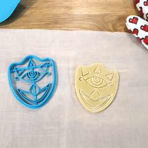 May include: A blue plastic cookie cutter and a beige cookie in the shape of a stylized eye with triangles and a crescent moon. Both are on a white parchment paper.
