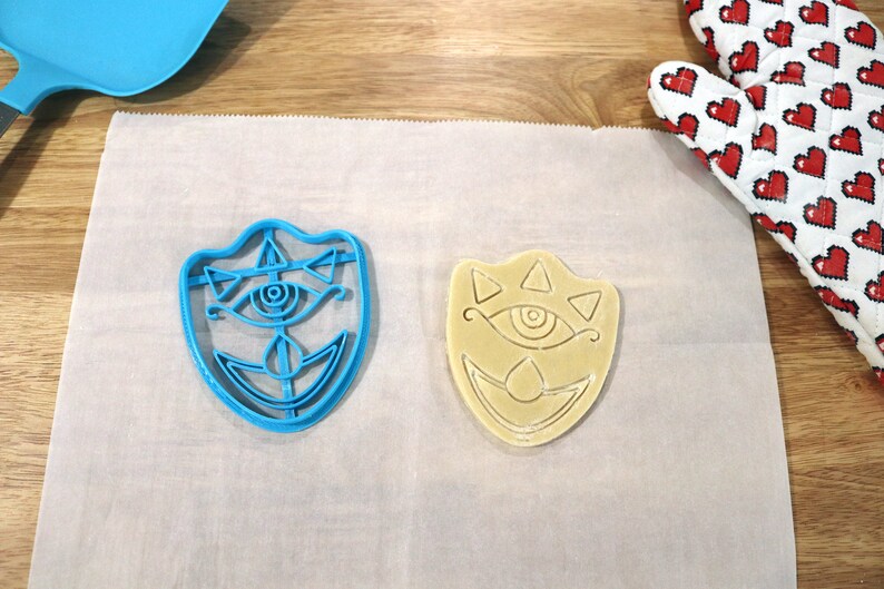 May include: A blue plastic cookie cutter and a beige cookie in the shape of a stylized eye with triangles and a crescent moon. Both are on a white parchment paper.