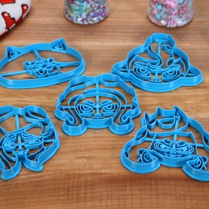 May include: Five blue plastic cookie cutters shaped like cartoon faces. The cutters are on a wooden surface.