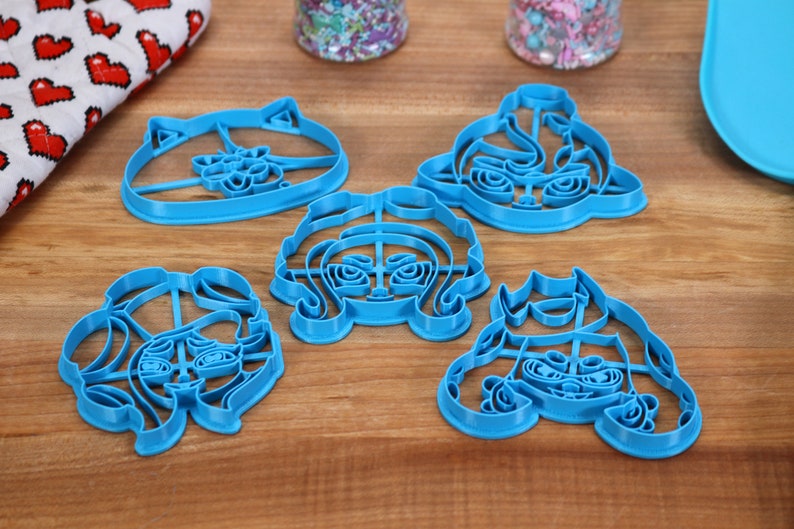 May include: Five blue plastic cookie cutters shaped like cartoon faces. The cutters are on a wooden surface.