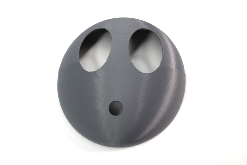 May include: A gray plastic face mask with two large eye holes and a smaller hole in the center. The mask is designed to resemble a face.