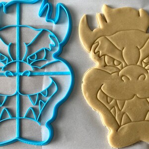 Op de afbeelding: Een blauwe plastic koekjesvormer in de vorm van een cartoonfiguur met een dreigende uitdrukking, naast een koekje dat met de vormer is uitgestoken.