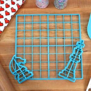 May include: Blue plastic cookie cutters shaped like a chess king and knight, a blue plastic grid, a wooden rolling pin, a blue spatula, and a red and white oven mitt with a heart pattern.