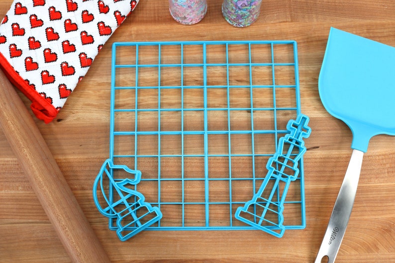 May include: Blue plastic cookie cutters shaped like a chess king and knight, a blue plastic grid, a wooden rolling pin, a blue spatula, and a red and white oven mitt with a heart pattern.