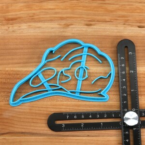 May include: A blue plastic cookie cutter shaped like a turkey. The cutter is 3 inches wide and 2 inches tall. It is sitting on a wooden surface.