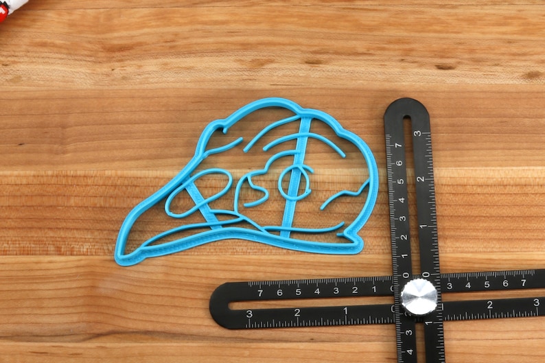 May include: A blue plastic cookie cutter shaped like a turkey. The cutter is 3 inches wide and 2 inches tall. It is sitting on a wooden surface.