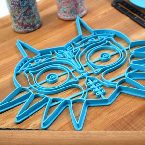 May include: A blue plastic cookie cutter shaped like the iconic Majora's Mask from the Legend of Zelda video game series. The cutter is detailed with the mask's signature features, including the eyes, nose, and horns.