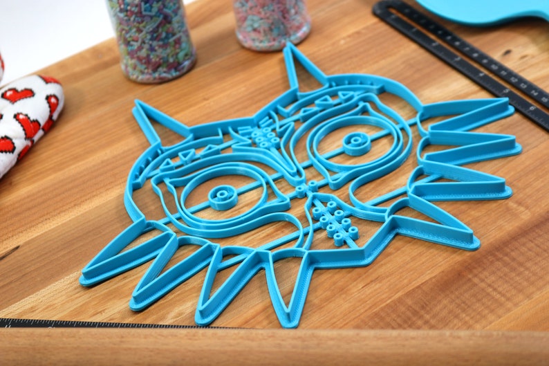 May include: A blue plastic cookie cutter shaped like the iconic Majora's Mask from the Legend of Zelda video game series. The cutter is detailed with the mask's signature features, including the eyes, nose, and horns.