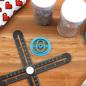 May include: A blue plastic cookie cutter shaped like a donut with a diamond in the center, on a black ruler with white markings. The ruler is on a wooden surface.