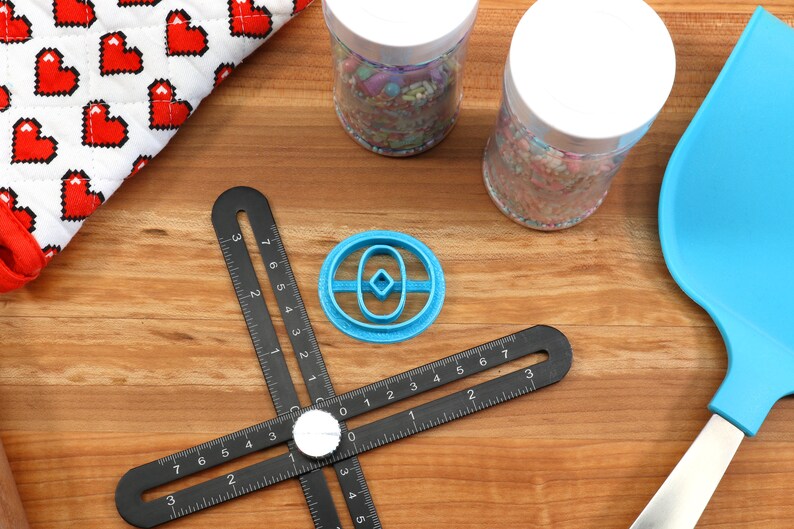 May include: A blue plastic cookie cutter shaped like a donut with a diamond in the center, on a black ruler with white markings. The ruler is on a wooden surface.