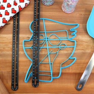 May include: A blue plastic cookie cutter shaped like a cartoon character, a wooden rolling pin, a red and white oven mitt, two black metal rulers, a blue silicone spatula with a silver handle, and two jars of sprinkles on a wooden surface.