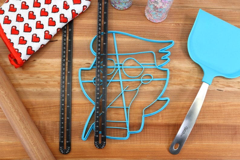 May include: A blue plastic cookie cutter shaped like a cartoon character, a wooden rolling pin, a red and white oven mitt, two black metal rulers, a blue silicone spatula with a silver handle, and two jars of sprinkles on a wooden surface.