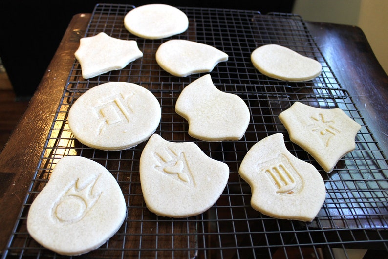 May include: A close-up of ten white sugar cookies on a wire rack. The cookies are decorated with various symbols, including a star, a circle with a symbol inside, and a shield with a symbol inside.
