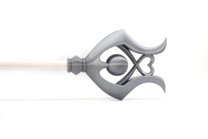 May include: A silver metal curtain rod finial with a decorative design featuring a heart and a circle. The finial is attached to a white curtain rod.
