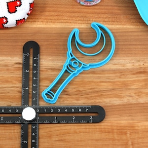 May include: Blue plastic cookie cutter shaped like a crescent moon wand. The wand has a handle and a crescent moon shape with a smaller crescent moon inside.