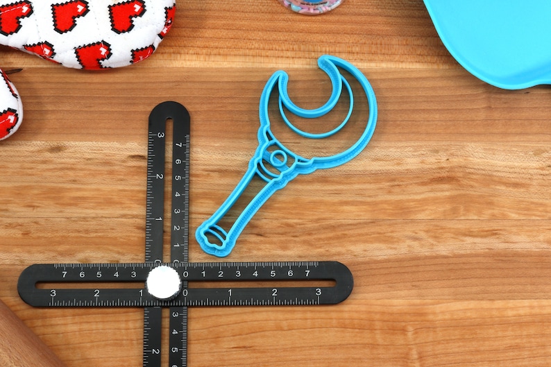 May include: Blue plastic cookie cutter shaped like a crescent moon wand. The wand has a handle and a crescent moon shape with a smaller crescent moon inside.