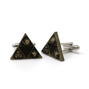 May include: A pair of gold-toned cufflinks featuring a triangular design with a black background. The triangle has four symbols on it, including a bird, a star, a crescent moon, and a stylized animal head.