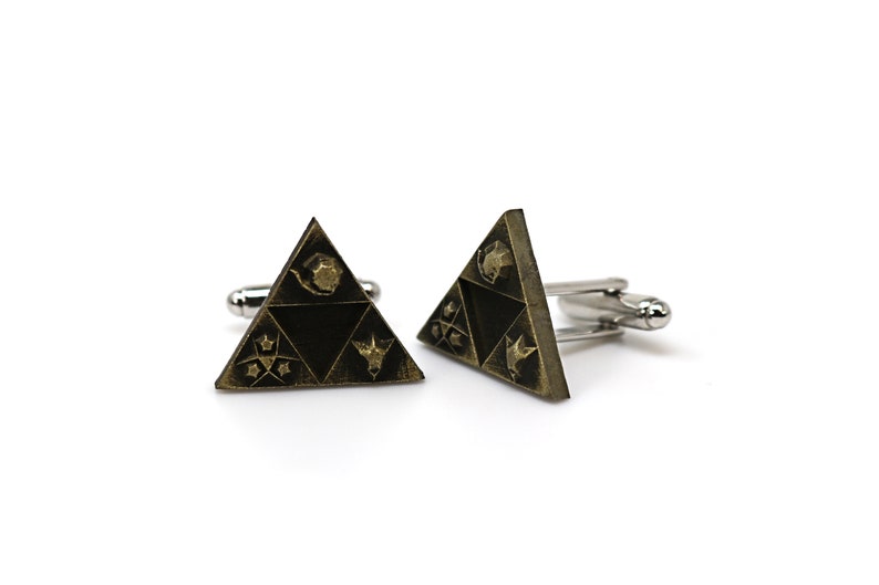 May include: A pair of gold-toned cufflinks featuring a triangular design with a black background. The triangle has four symbols on it, including a bird, a star, a crescent moon, and a stylized animal head.