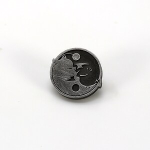 May include: A silver-toned yin yang pin with a cartoon dragon and a cartoon cat design.