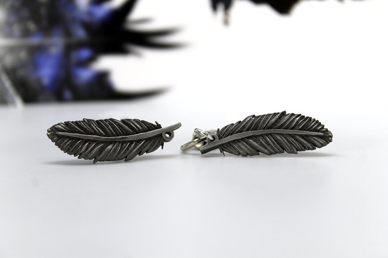 Op de afbeelding: Twee zilverkleurige veervormige bedels. Elke bedel heeft gedetailleerde veeradermotieven. De bedels zijn verbonden door een kleine ring. De achtergrond is wit met vage blauwe en zwarte elementen.