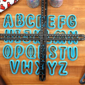 May include: A set of 26 blue plastic alphabet cookie cutters on a wooden surface. The cutters are arranged in alphabetical order and are centered on a black ruler.