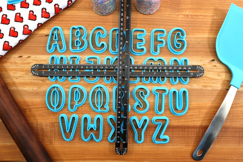 May include: A set of 26 blue plastic alphabet cookie cutters on a wooden surface. The cutters are arranged in alphabetical order and are centered on a black ruler.