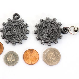 May include: A silver metal keychain with a circular design featuring a stylized symbol and a silver metal clasp. The keychain is surrounded by four coins.