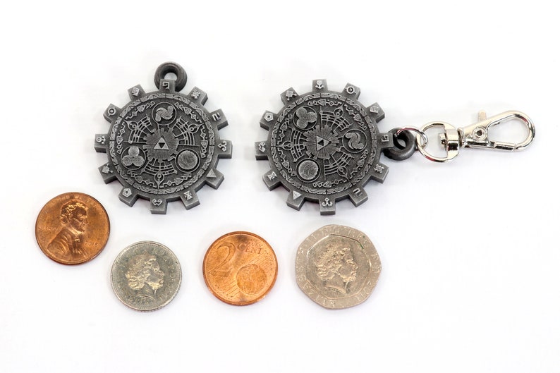 May include: A silver metal keychain with a circular design featuring a stylized symbol and a silver metal clasp. The keychain is surrounded by four coins.