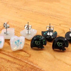 May include: Four pairs of earrings with different designs. Two pairs are white with a blue design and two pairs are black with a green design. The earrings are made of resin and have a glittery finish.