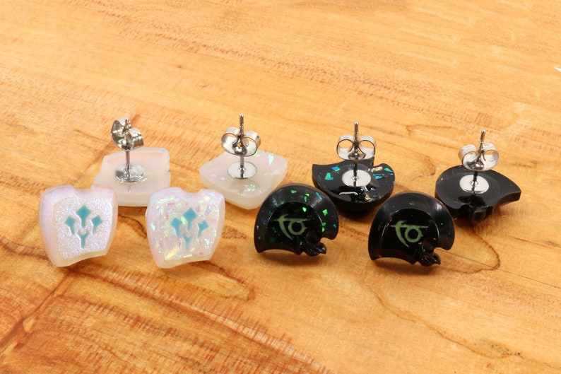 May include: Four pairs of earrings with different designs. Two pairs are white with a blue design and two pairs are black with a green design. The earrings are made of resin and have a glittery finish.