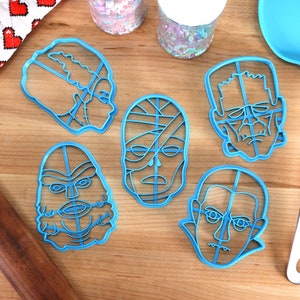 May include: A collection of six blue plastic cookie cutters in the shapes of classic movie monsters, including Frankenstein's monster, the Mummy, and Dracula. A blue spatula and a rolling pin are also visible on a wooden surface.