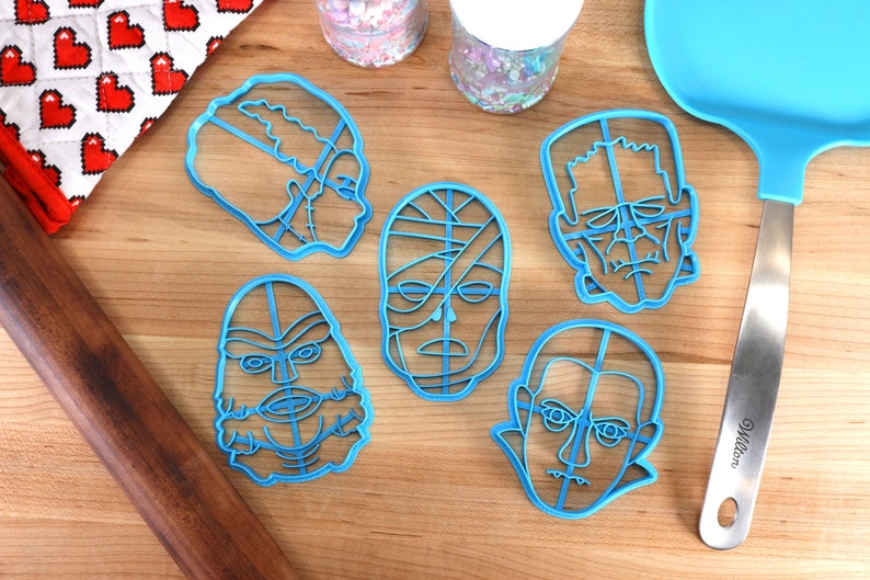 May include: A collection of six blue plastic cookie cutters in the shapes of classic movie monsters, including Frankenstein's monster, the Mummy, and Dracula. A blue spatula and a rolling pin are also visible on a wooden surface.