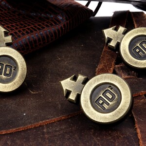 May include: Three gold-colored metal tokens with a symbol and the letters "AD" with a superscript "2" on each. The tokens are on a brown leather surface.